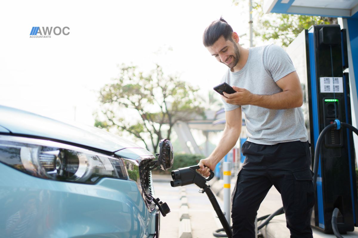 Man charging electric car at EV charging station while checking smartphone – concept of eco-friendly driving and tax benefits