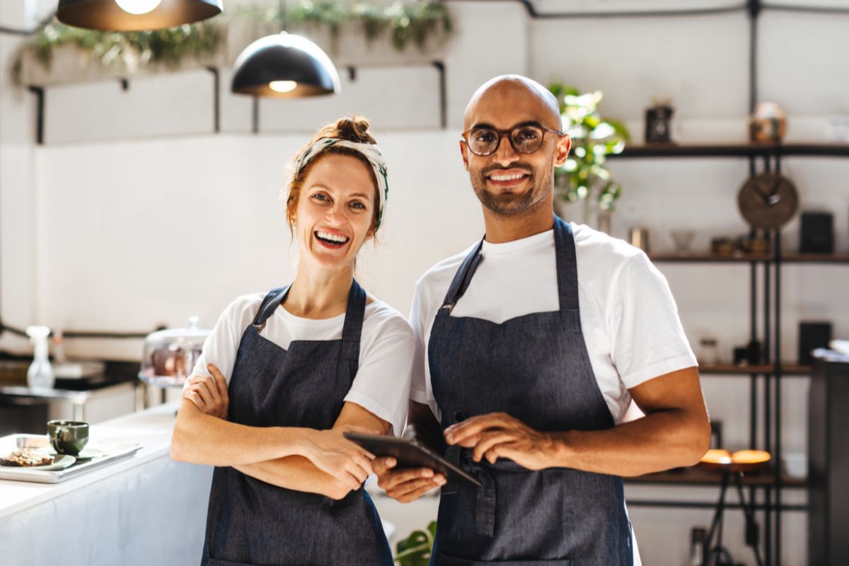 Two small business owners smiling in their café, wearing aprons and holding a tablet – representing small businesses affected by National Insurance changes in 2025